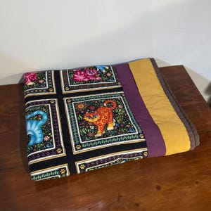 May include: A vibrant quilt with a repeating pattern of cartoon cats in blue, orange, and pink on a black background. The quilt has a purple and gold border, with a brown edge.