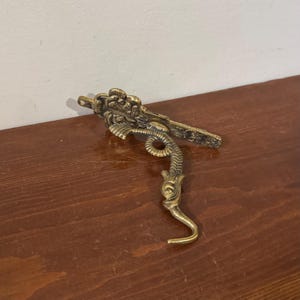 May include: A brass dragon-shaped hook with detailed features. The hook has a dragon's head, body, and tail, with a curved hook at the end. The dragon is a golden colour and appears to be made of metal. The hook is designed to be mounted on a wall or other surface.
