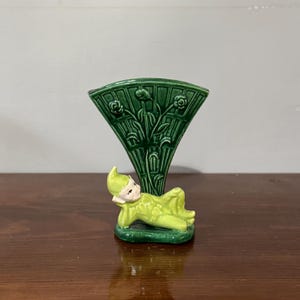 May include: A green ceramic vase with a whimsical design featuring a reclining elf figure and a stylised floral motif. The vase is perfect for adding a touch of whimsy to any home decor.