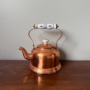 May include: A polished copper tea kettle featuring a rounded body and a curved spout. The kettle includes a white ceramic handle and a matching lid knob, both decorated with small blue cross-hatch patterns. The metal surface reflects light.