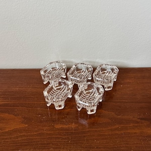 May include: A set of five clear glass knife rests, each featuring a rectangular shape with four small feet and a textured, ribbed surface pattern on the top. These small, transparent glass pieces are arranged in a cluster on a flat surface.