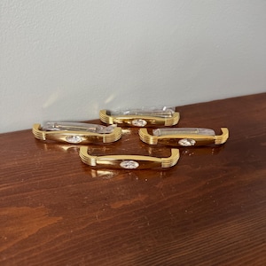 May include: Four gold-toned metal cabinet pulls, each featuring a central oval-shaped clear crystal accent and ribbed detailing on the ends. The hardware pieces are individually wrapped in clear plastic packaging with included mounting screws.