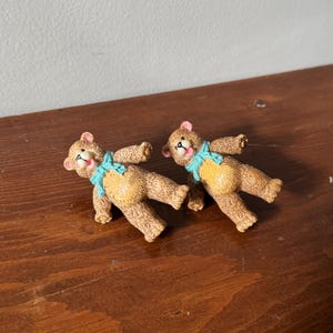 May include: Two brown teddy bear figurines with blue ribbon bows. The bears are lying on their backs with their paws in the air.