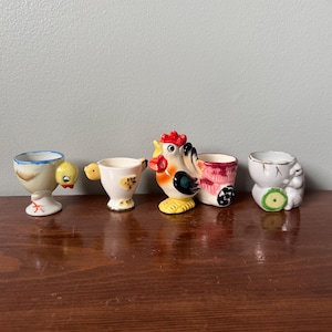 May include: A set of four ceramic egg cups featuring animal designs. The first is a bird, the second a small chick, the third a rooster pulling a cart, and the fourth a white animal with green accents. Each cup is painted in vibrant colors.
