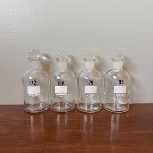 May include: Four clear glass apothecary bottles with ground glass stoppers. Two bottles are labelled Wheaton U.S.A. with numbers 172 and 128. Two bottles are labelled Vitro U.S.A. with numbers 79 and 85. Each bottle features a white rectangular label area.