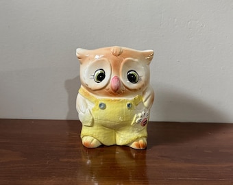 May include: A ceramic owl figurine with large, expressive eyes and a pink beak. The owl is wearing yellow overalls with a small pocket. The owl's head is a light orange color, and the body is white.