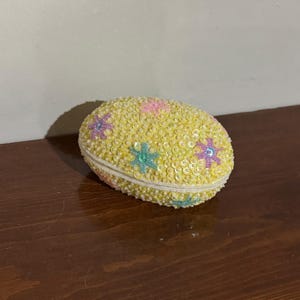 May include: A yellow, oval trinket box covered in sequins. The box is decorated with colourful star-shaped sequins in pink, purple, and blue. The edges are trimmed with a white beaded border. The box is closed and sits on a dark wooden surface.