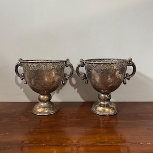 Peut inclure: Deux gobelets anciens de couleur argentée avec des poignées ornées et des bords décoratifs. Les gobelets ont un socle et présentent des signes d'âge et d'usure, suggérant qu'il s'agit de pièces vintage ou antiques. Ils sont exposés sur une surface en bois.
