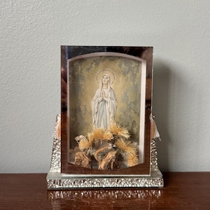 May include: A rectangular shadow box frame with a mirrored border and a textured silver base. Inside, a print of the Virgin Mary in a white and blue robe stands above the text JE SUIS L'IMMACULÉE CONCEPTION, accented by dried golden pine needles.
