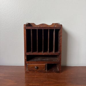 May include: A dark brown wooden wall-mounted organizer featuring five vertical slots for documents, a horizontal shelf, one small pull-out drawer with a round knob, and an open compartment next to the drawer for additional storage space.