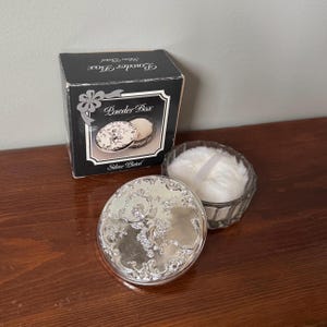 May include: A silver-plated powder box with a round lid featuring an embossed cherub and floral design. The set includes a clear glass base containing a white fluffy powder puff and a black cardboard box labeled Powder Box Silver Plated.