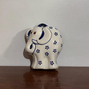 Pode incluir: Uma estatueta de elefante de cerâmica branca com detalhes florais azuis e uma argola azul escura para pendurar. O elefante tem olhos castanhos e detalhes azuis nas orelhas e na tromba. A estatueta está numa superfície castanha.