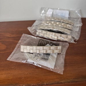 May include: A set of five silver-toned metal cabinet pulls, each featuring a textured basket-weave pattern. One pull is displayed outside of its individual clear plastic packaging, while the other four remain sealed in their original plastic bags.