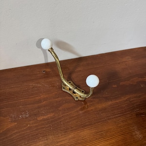 Peut inclure: Une patère murale en laiton poli dotée de deux bras incurvés. Chaque bras se termine par un bouton en céramique blanche, lisse et rond. La plaque de base est rectangulaire avec des coins arrondis et comporte quatre trous de fixation.