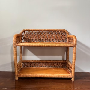 May include: A two-tiered light brown rattan shelf featuring a woven lattice pattern on the back and side panels. The unit has two flat, smooth shelves supported by a sturdy frame constructed from curved rattan poles with a natural finish.
