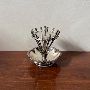 May include: A silver-plated hors d'oeuvre pick holder. It features a scalloped dish base, a central stem, and a circular arrangement of picks. Each pick has a decorative top, creating a tiered design. A vintage serving piece.