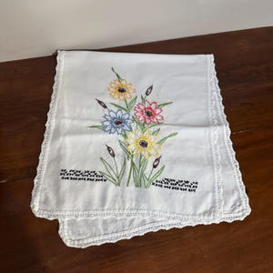 Peut inclure: Chemin de table brodé blanc avec un motif floral. Les fleurs sont jaunes, roses et bleues, avec des feuilles vertes et des quenouilles brunes. Les bords sont finis avec une délicate bordure en dentelle.