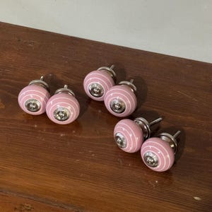 Puede incluir: Seis pomos de armario rosas y blancos a rayas con herrajes plateados. Los pomos son redondos con un centro plateado decorativo. Los pomos están dispuestos sobre una superficie de madera.