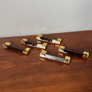 May include: A set of four drawer pulls featuring a long, dark, textured rectangular centre bar flanked by two gold-toned metal ends. Each end piece has a stepped, ribbed design on the outer edges, providing a contrast between the dark finish and the bright, reflective gold metal components.