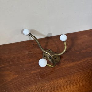 May include: A wall-mounted coat hook featuring a brass-toned metal frame with a textured mounting plate and three curved arms. Each arm terminates in a smooth, round white ceramic knob, providing three distinct points for hanging items.