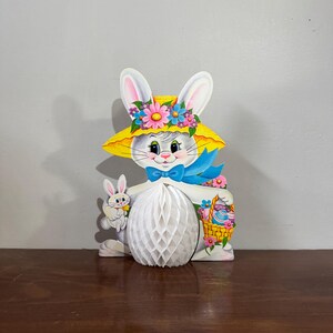 May include: A decorative Easter bunny wearing a yellow hat decorated with flowers. The bunny holds a small bunny and a basket of Easter eggs. The bunny's body is a white honeycomb paper design. A blue bow tie adds colour to the design.