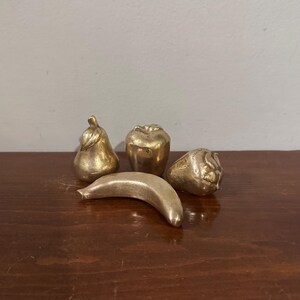 May include: A collection of four metallic gold-toned decorative fruit figurines. The set includes a pear, an apple, a banana, and a strawberry. Each piece features a smooth, reflective surface with subtle contours that mimic the natural shapes of fruit.