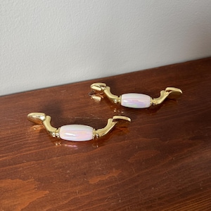 May include: Two cabinet pulls featuring a polished gold-tone metal finish with a central, cylindrical, iridescent white insert. Each handle has a curved, ergonomic shape with mounting points at both ends, showcasing a reflective, pearlescent surface.