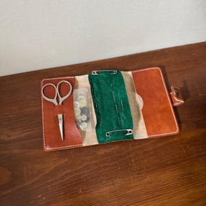May include: Open leather sewing kit with scissors, a needle threader, and a small plastic bag of buttons. The kit also includes a green felt section with needles secured by safety pins. The kit is brown with a tan and orange striped fabric.