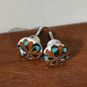 May include: Two round ceramic cabinet knobs featuring a hand-painted floral pattern with brown, black, and bright blue petals on a white base. Each knob has a silver-toned metal screw extending from the back for installation purposes.
