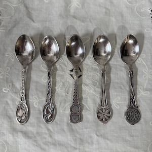 May include: Five antique silver spoons with decorative handles. Each spoon features a unique design, such as floral motifs, scrollwork, and geometric patterns. The spoons are displayed in a row on a white, textured surface.