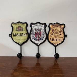 May include: Three Absinthe-themed wall hooks. Each hook has a shield-shaped design with vintage graphics and text, including "ABSINTHE" and French text. The hooks are black with a round knob at the bottom.