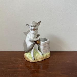 May include: A vintage porcelain figurine of a cat-like creature holding a small barrel, next to a miniature well. The figurine is primarily white and gray, with a touch of brown and green. The well is painted with pink and white bricks.