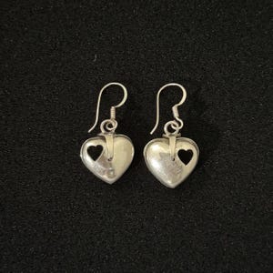 May include: A pair of silver heart-shaped dangle earrings. Each earring features a heart cutout and a curved hook for pierced ears. The earrings are displayed on a black surface.