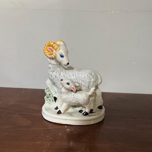 May include: A ceramic figurine of a ram and a lamb. The ram has large, curled horns with yellow and pink accents. The lamb stands beside the ram. Both have blue eyes and are on a white base with green accents.