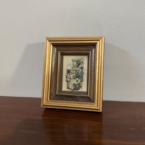 May include: A framed print featuring a black and white illustration of a group of people. The frame is a combination of gold and brown, with a detailed border. The artwork is set on a dark wooden surface.
