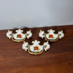 May include: Three white ceramic drawer pulls with gold-toned metal edges and swinging handles. Each pull features a central floral decal depicting a red rose with green leaves. The hardware has a decorative, scalloped shape and gold accents.
