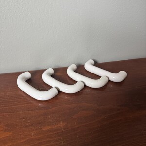 Peut inclure: Quatre poignées de tiroir en céramique blanche avec une finition lisse et brillante. Chaque poignée présente un design simple en forme de U incurvé avec deux points de fixation aux extrémités, disposées en rangée pour montrer leur forme arrondie.