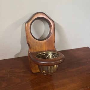 May include: A wall-mounted wooden holder featuring a vertical back panel with a circular cutout at the top and a horizontal shelf section. A polished brass bowl is securely fitted into the circular opening of the wooden shelf, creating a functional unit.
