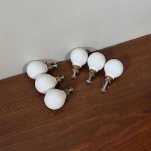 May include: Six white ceramic cabinet knobs with brass and silver fittings are arranged on a dark wood surface. The knobs are round and bulbous, with a threaded screw for attachment. Each knob is approximately 3 cm in diameter.