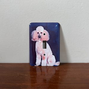 Puede incluir: Interruptor de luz con una ilustración de un caniche rosa claro sobre un fondo azul oscuro. El caniche está representado sentado. El interruptor es rectangular con esquinas redondeadas.
