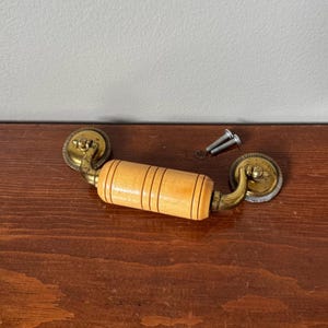 May include: A furniture pull featuring a cylindrical light-coloured wooden handle with three horizontal grooves. The handle is attached to two circular brass mounting plates with decorative scalloped edges. Two small metal screws are included.