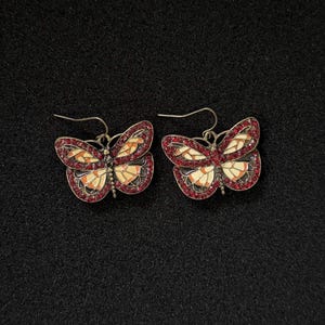 May include: A pair of butterfly earrings with red and orange accents. The earrings have a silver-tone metal finish and are adorned with small, red gemstones around the edges of the butterfly wings. The wings feature a cream, orange, and black design.