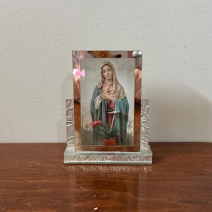 Puede incluir: Imagen religiosa de una mujer con una túnica azul y verde, enmarcada en un rectángulo de vidrio transparente con una base decorativa. La imagen presenta a una mujer con un halo, un corazón rojo y una pequeña planta en maceta.