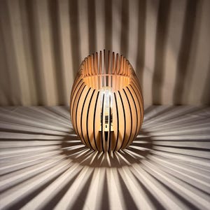 Könnte beinhalten: Holz-Tischlampe mit einzigartigem Design. Die Lampe hat einen hellbraunen, oval geformten Schirm aus vertikalen Holzlatten. Die Lampe wirft einen gestreiften Schatten auf die darunter liegende Oberfläche. Die Glühbirne ist im Inneren des Schirms sichtbar.
