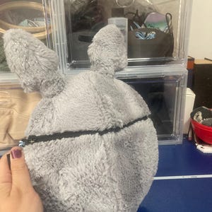 Op de afbeelding: Een grijze, pluche konijnvormige tas met lange oren en een zwarte ritssluiting. De tas wordt vastgehouden door een hand met een paars gelakte nagel. De achtergrond omvat doorzichtige plastic opbergdozen.