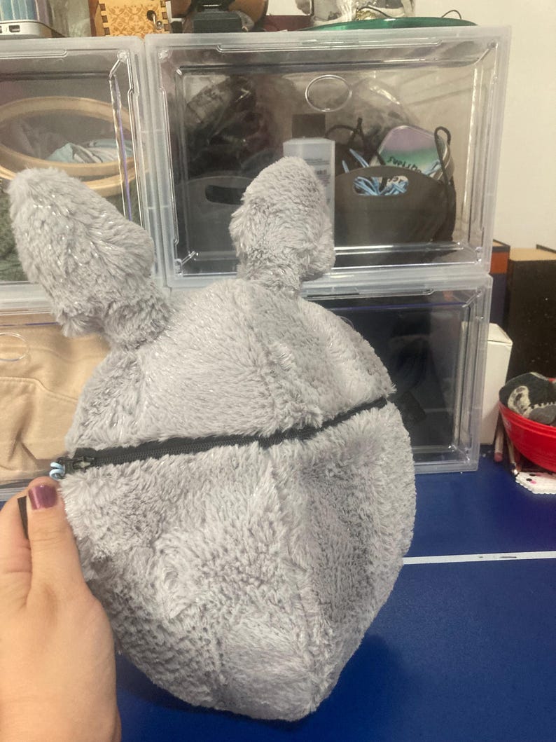 Op de afbeelding: Een grijze, pluche konijnvormige tas met lange oren en een zwarte ritssluiting. De tas wordt vastgehouden door een hand met een paars gelakte nagel. De achtergrond omvat doorzichtige plastic opbergdozen.