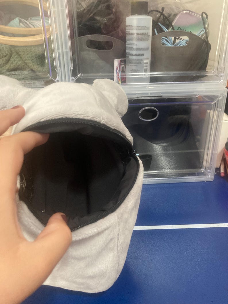 Op de afbeelding: Een lichtgrijze pluche beer-vormige pouch met een zwart interieur. De pouch is open, waardoor de binnenkant zichtbaar is. De rits is zwart. De pouch wordt voor een doorzichtige plastic opbergcontainer gehouden.