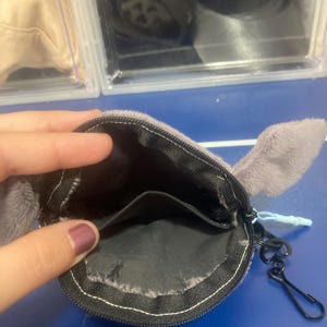 Op de afbeelding: Een grijze pluche munttas met een ritssluiting en twee kleine grijze oren. De binnenkant is zwart en er is een zwarte metalen sluiting bevestigd. De tas is open en onthult de binnenkant.