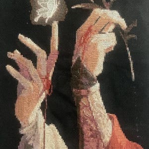 May include: A cross-stitch embroidery on a black fabric background depicting two hands. One hand reaches toward a white butterfly, while the other hand holds a pale flower. The design features shades of beige, red, and white thread on black cloth.