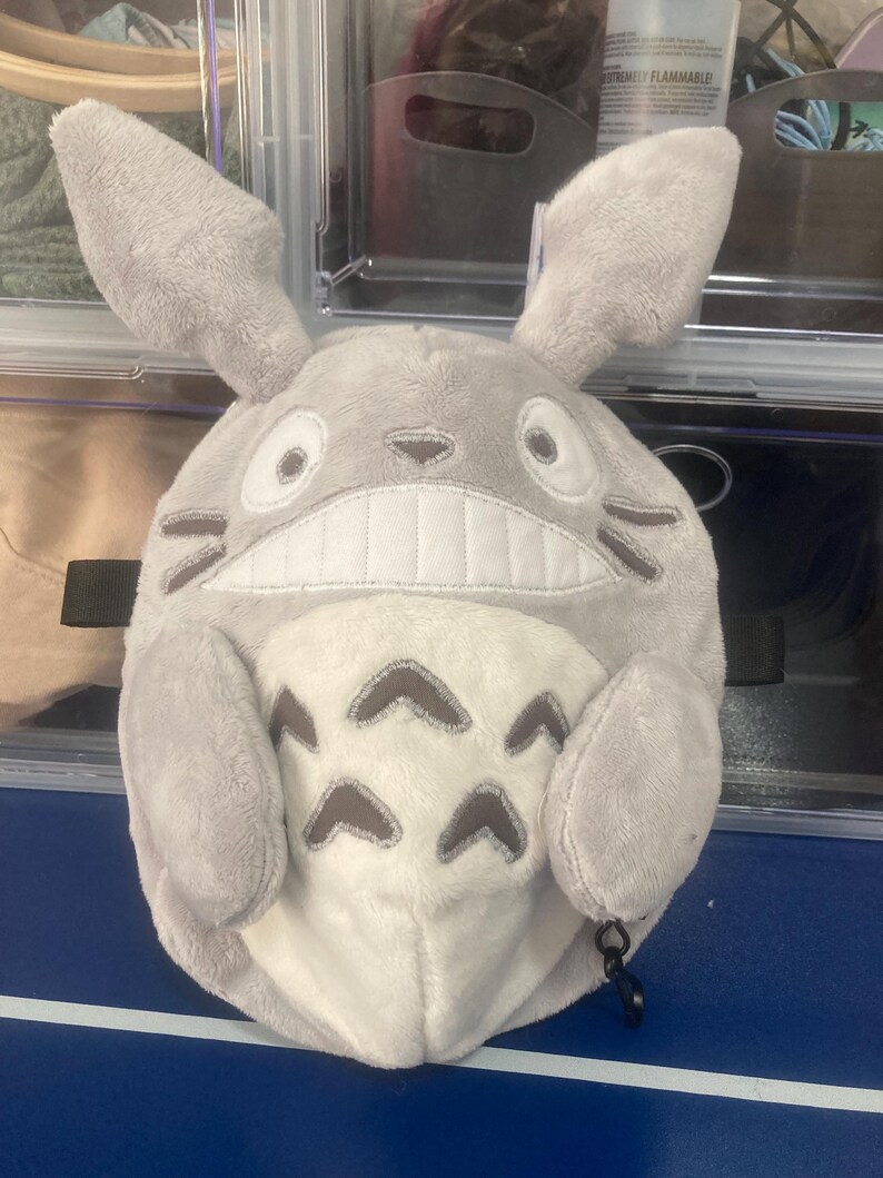 Op de afbeelding: Een pluche, grijze Totoro-ge&iuml;nspireerde karakter met grote oren, een brede grijns en donkergrijze markeringen. Het karakter heeft een witte buik met donkergrijze pijlvormige markeringen. Een zwarte clip is aan de onderkant bevestigd.